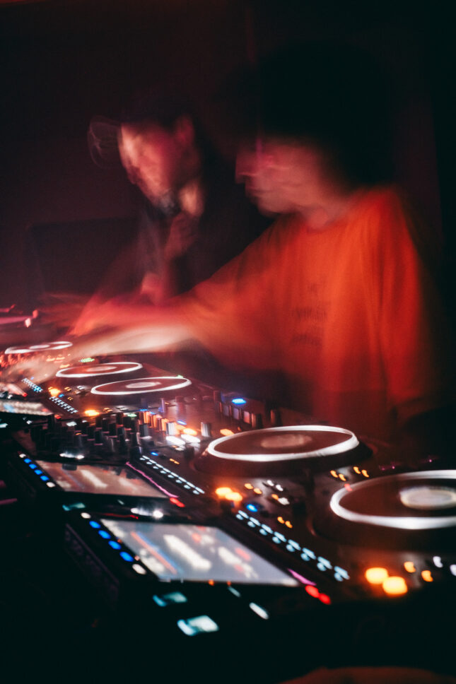 Long exposure image of two DJs behind the decks create ghostly figures as they move. Sustain-Release.