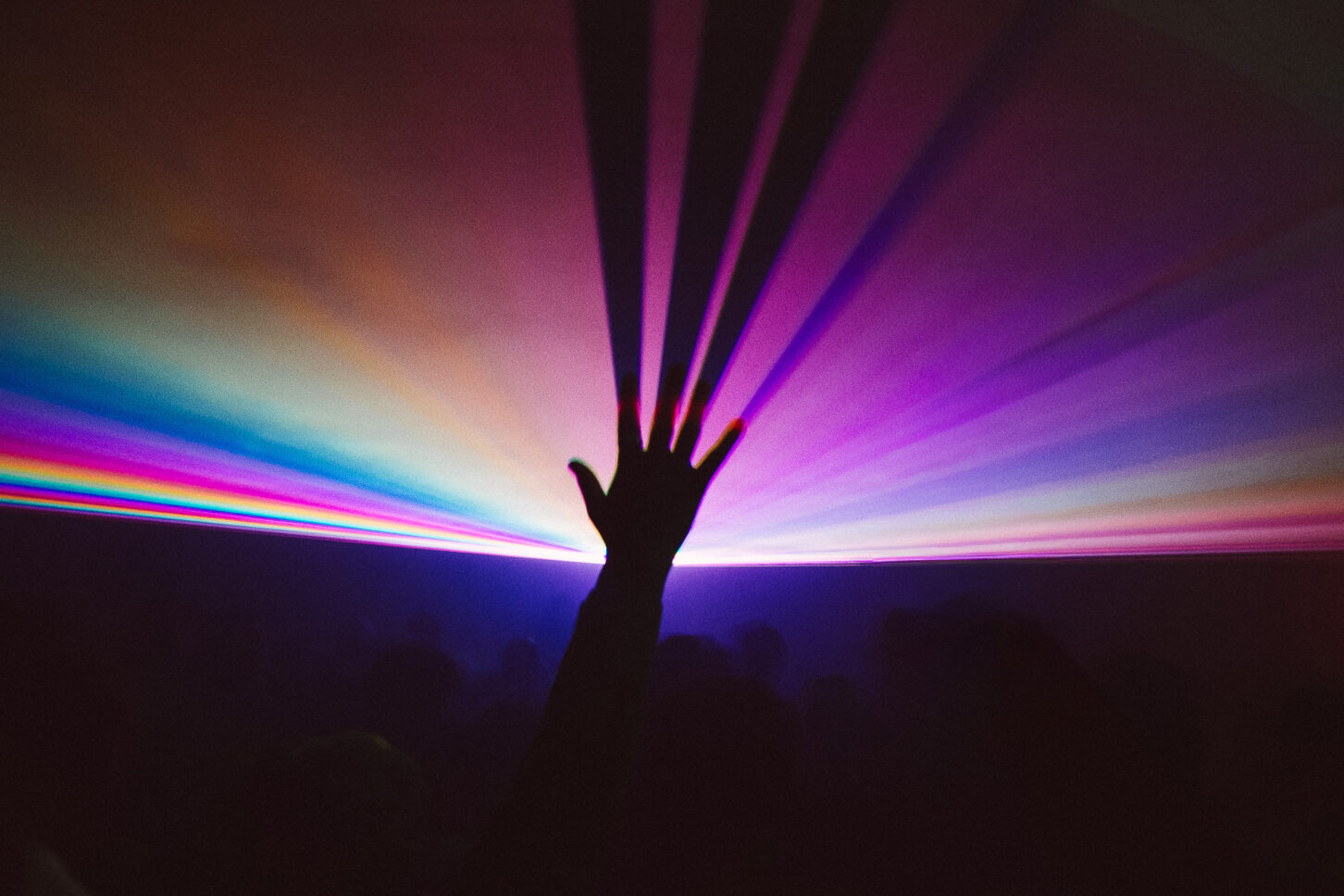 Silhouette of a hand reaches up high in a dim dance floor and touches a rainbow colored laser creating long streaking shadows where the fingers touch. Sustain-Release.