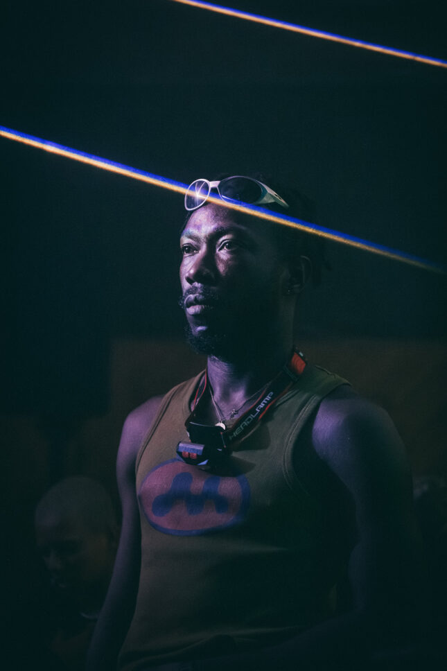 Man with 90's rave style sunglasses over his head and a camping headlamp stares blankly into space as laser lights from the dance floor intersect his head. Sustain-Release.