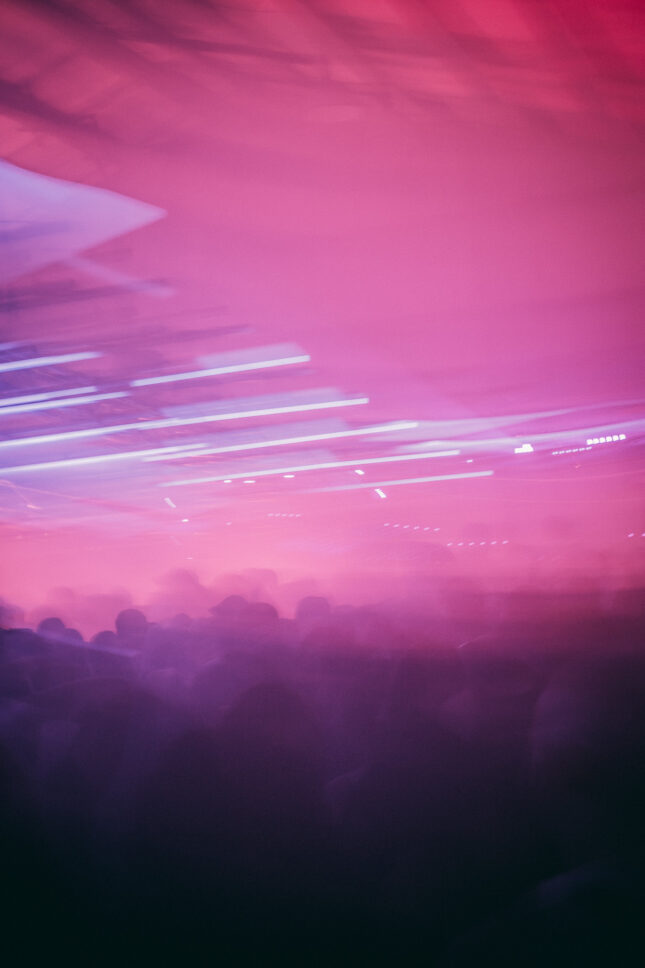 A long exposure image of Sustain-Release revelers under a pink light. The long exposure of the lights resemble a sandstorm sweeping over heir heads.