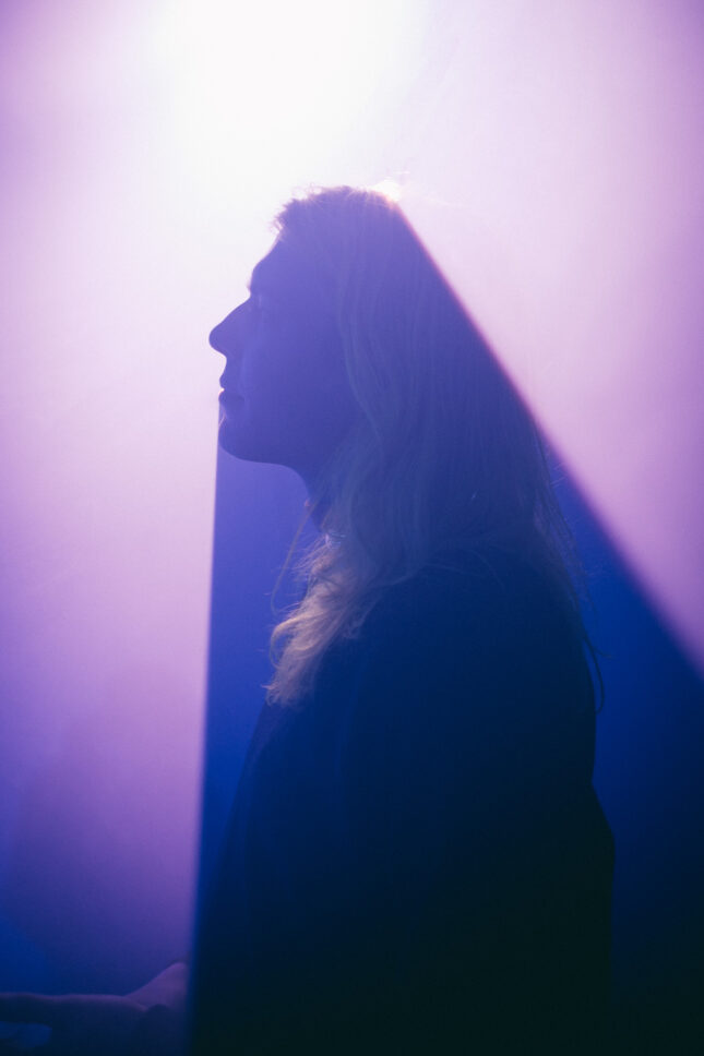 Man with neon green hair poses in front of purple light creating a shadow of his profile.
