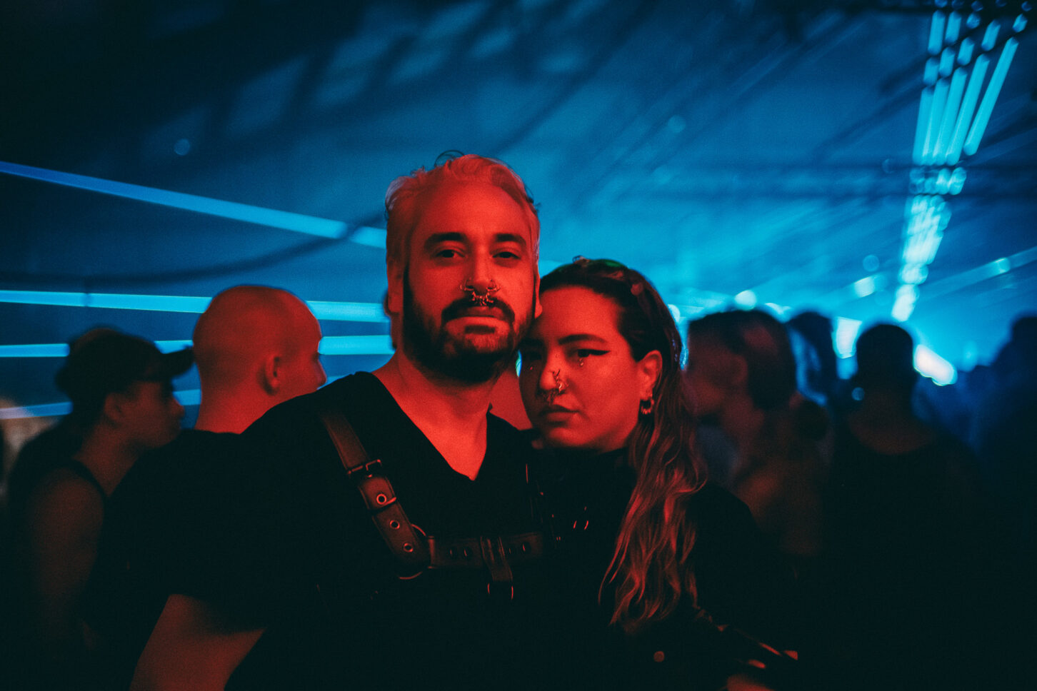 A couple wearing all black bondage outfits with nose piercings while covered in red light pose for the camera in the middle of the dance floor at Sustain-Release.