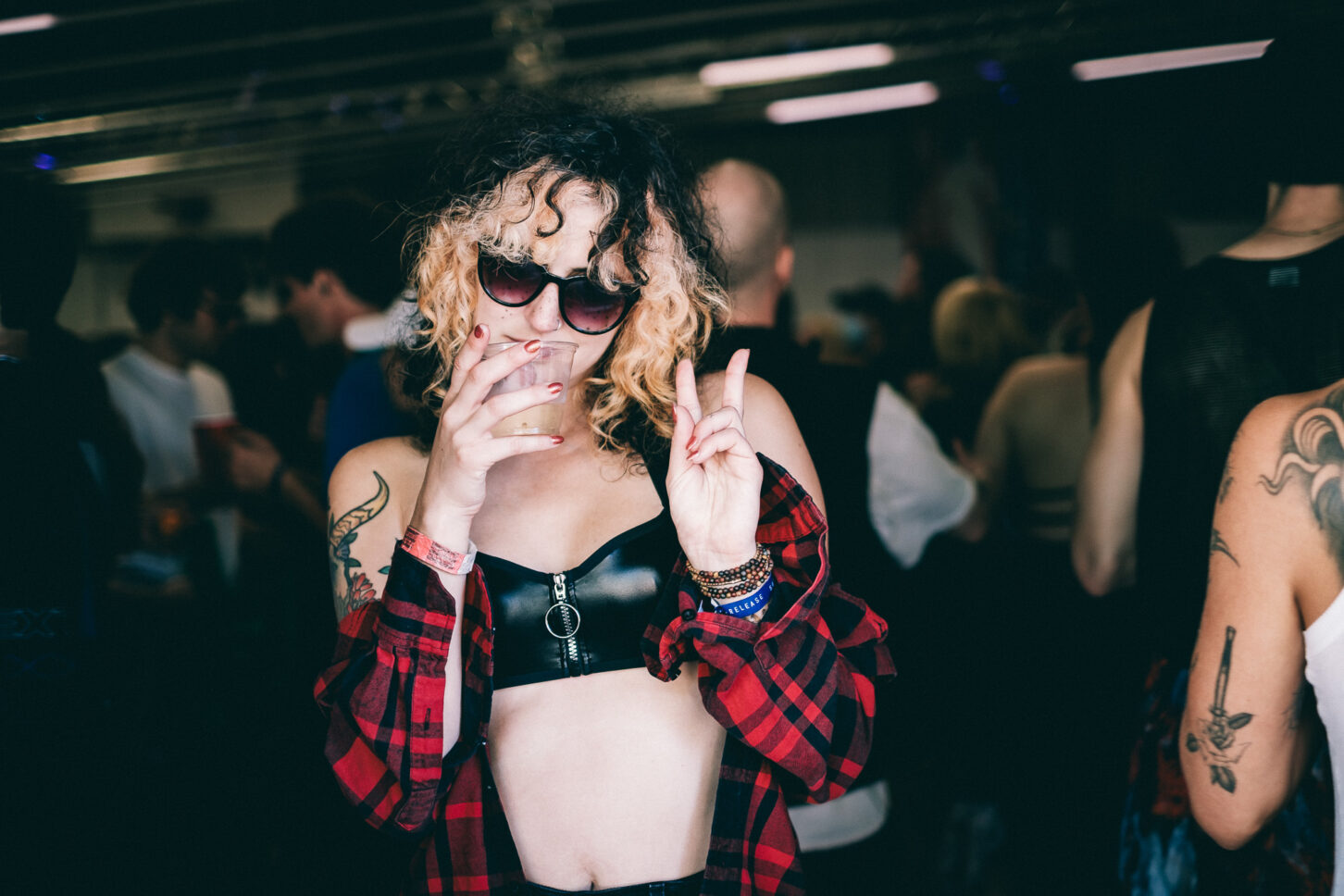 Girl with curly hair, tattoos, trendy sunglasses, red plaid shirt and leather bra holds a drink while showing a peace sign with her fingers.