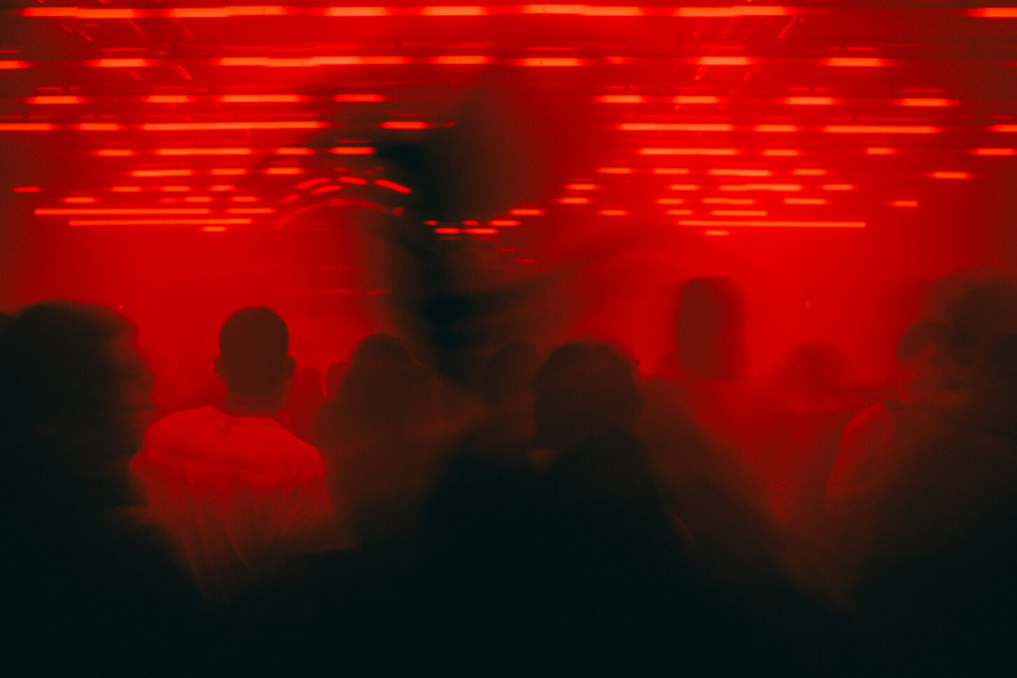 A long abstract image of a dance floor full of people moving with a person crossing in front of the camera and warping the lights with his silhouette.