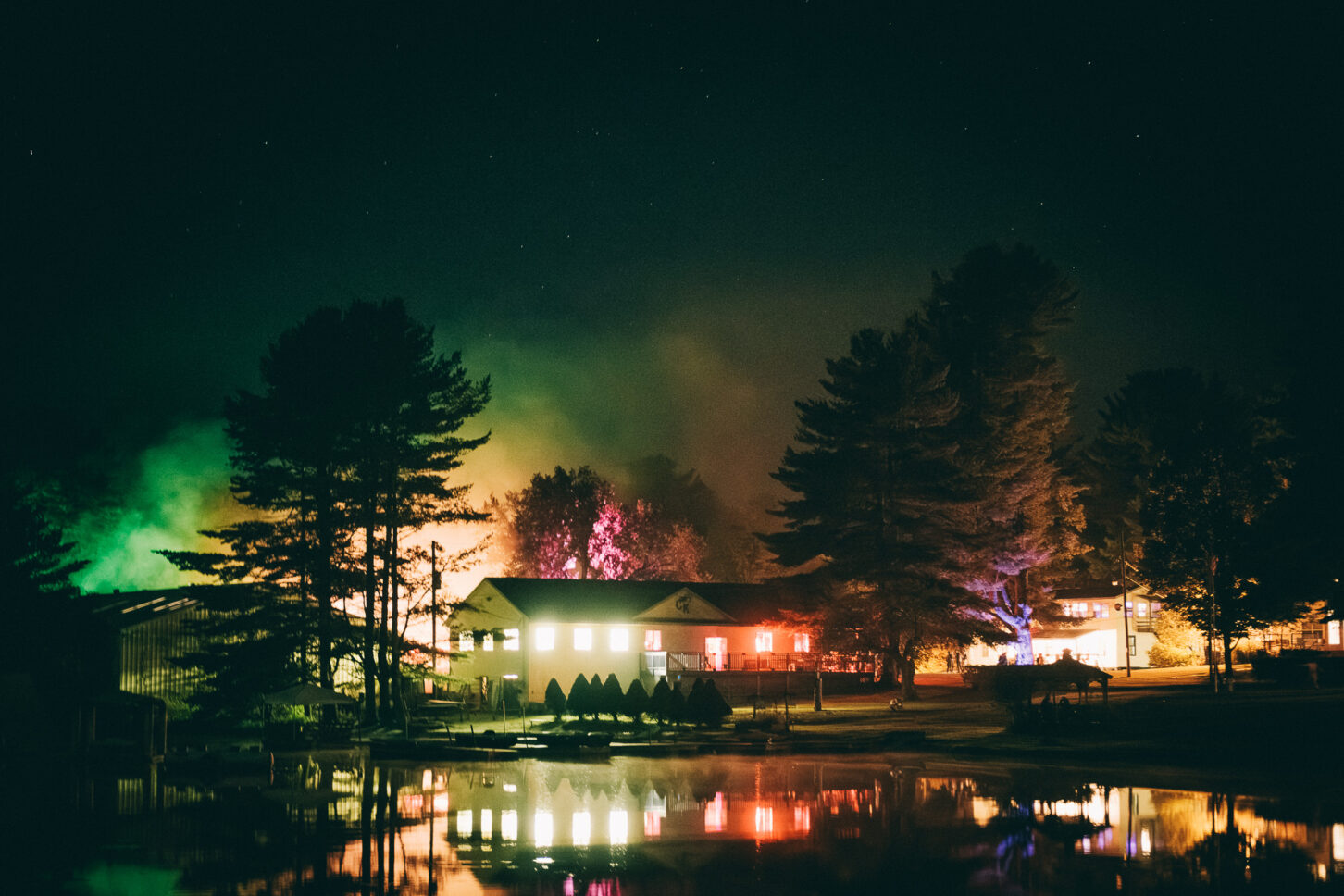 Night landscape photo of Camp Kennybrook's buildings across the lake under the stars during Sustain-Release. The area is covered with fog and neon lights emanate from the buildings.