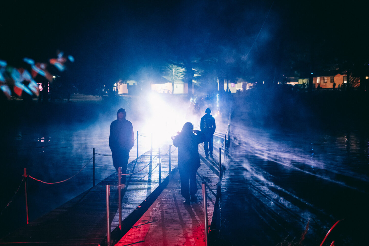 3 people walking across the wooden bridges across the lake in Camp Kennybrook at night during Sustain-Release.