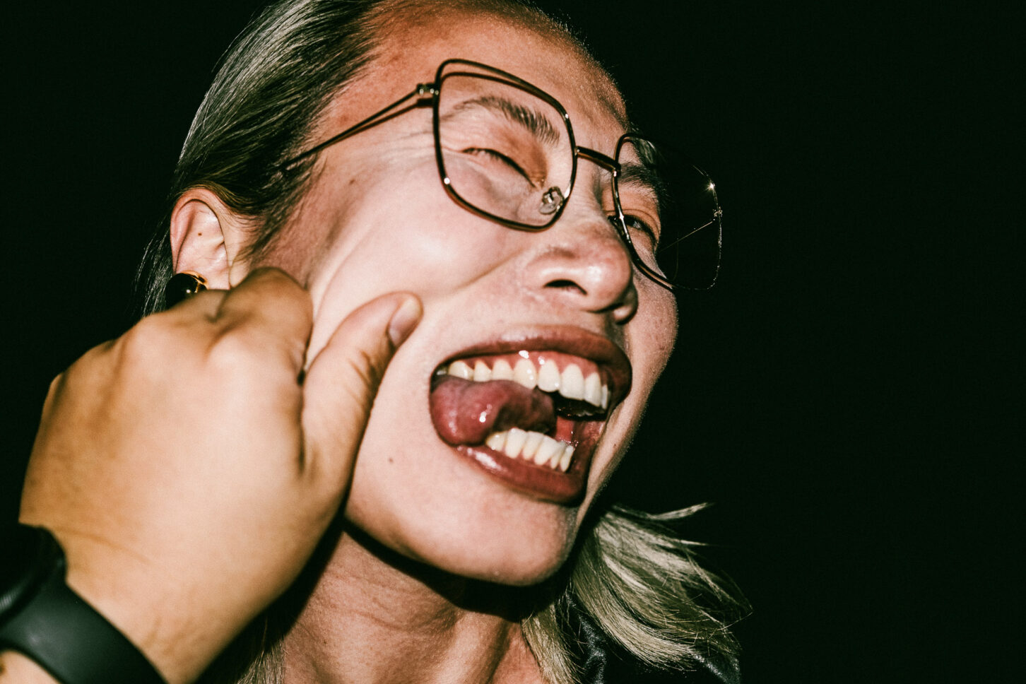 A person with a wide-eyed, shocked expression in a raw, gritty flash portrait from the 'Cheeky Bastards' series, reacting to a pinch on their left cheek.