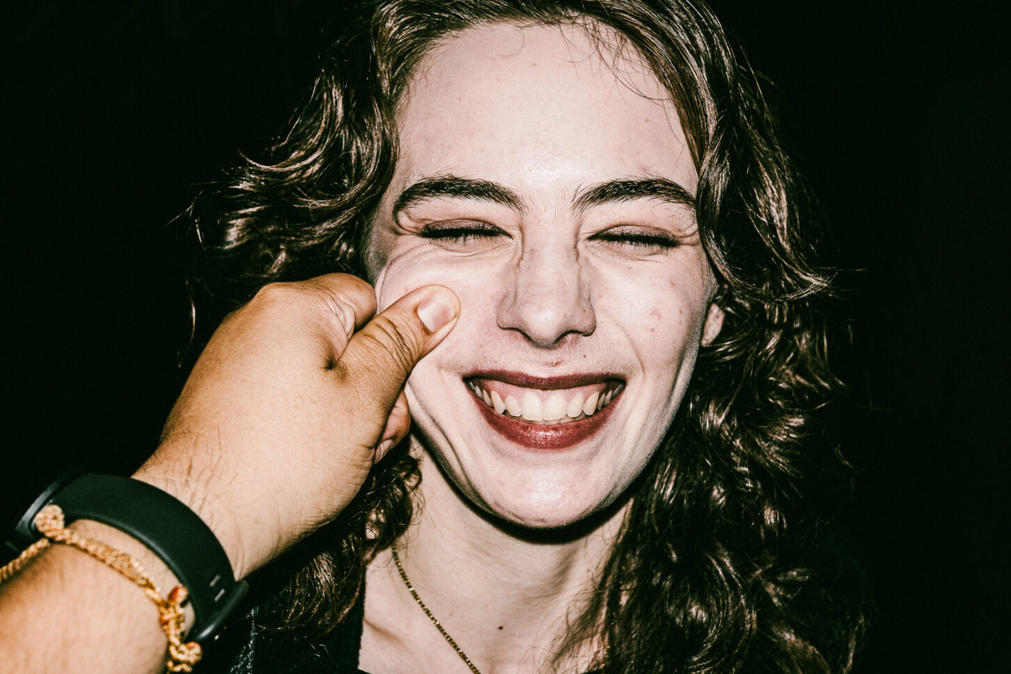 A woman with a playful, annoyed grimace captured with harsh flash lighting during a cheek-pinch for the 'Cheeky Bastards' series.
