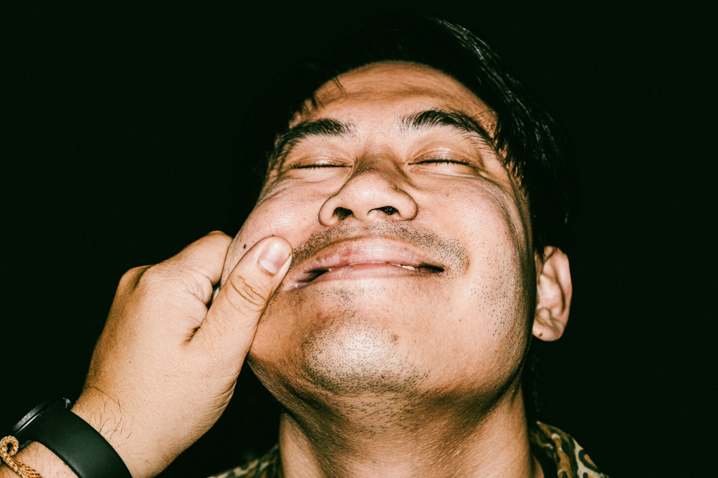 A person mid-laugh with eyes closed in a gritty, high-contrast flash photo, reacting to the signature left-cheek pinch of the 'Cheeky Bastards' series.