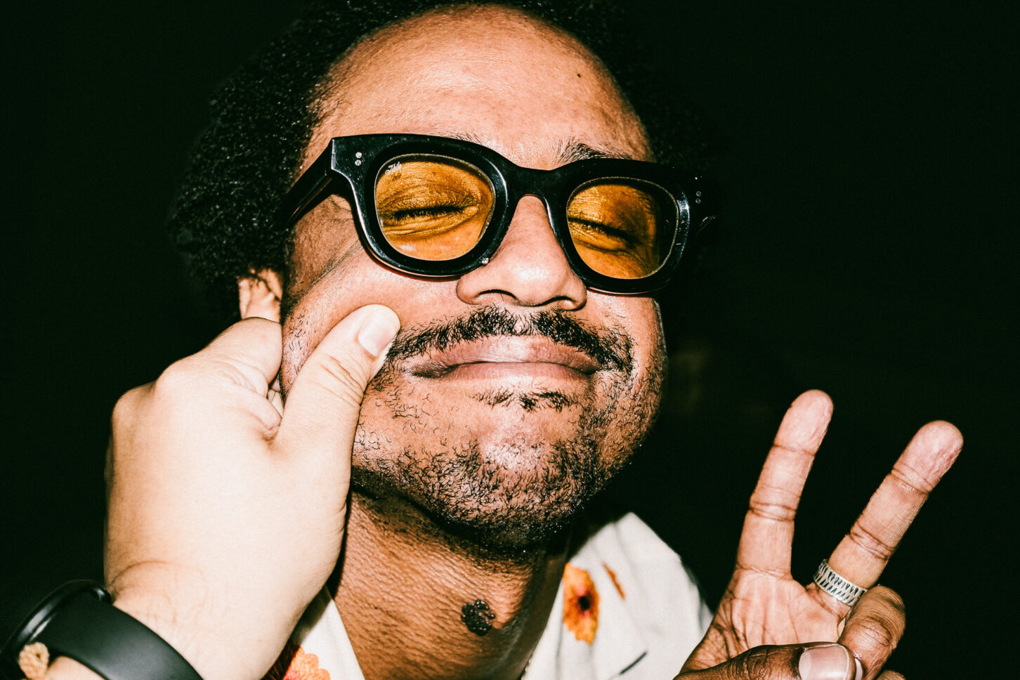 A man with a mischievous, squinted grin in a gritty street-style flash portrait, part of the 'Cheeky Bastards' photography project.