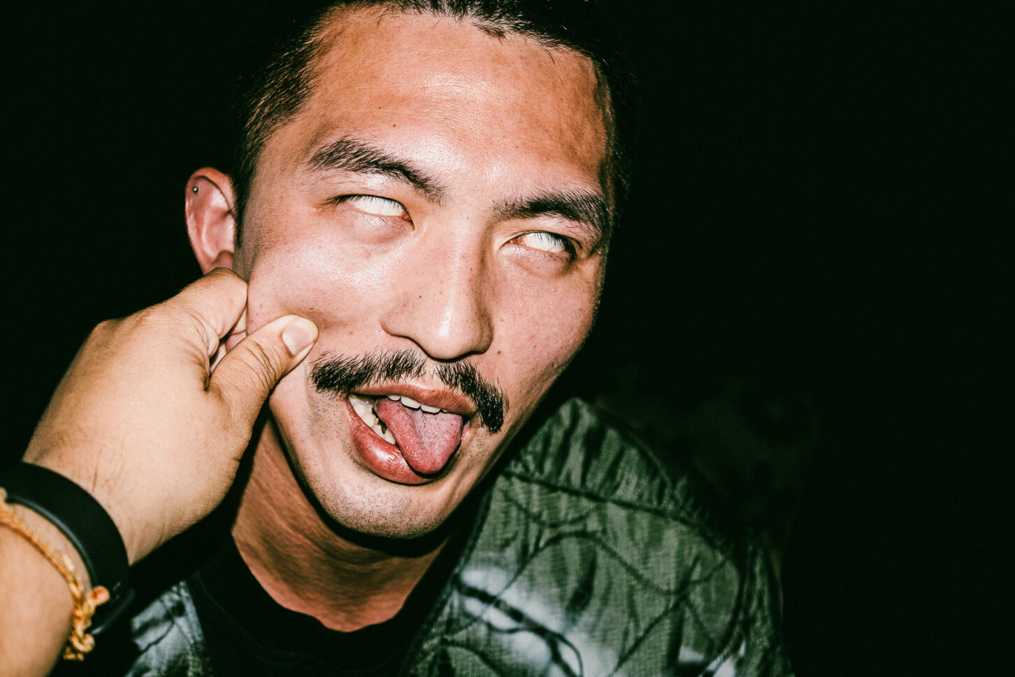 A person rolling his eyes with his tongue out in a candid, harsh-light photo from the 'Cheeky Bastards' series, reacting to the signature cheek pinch.