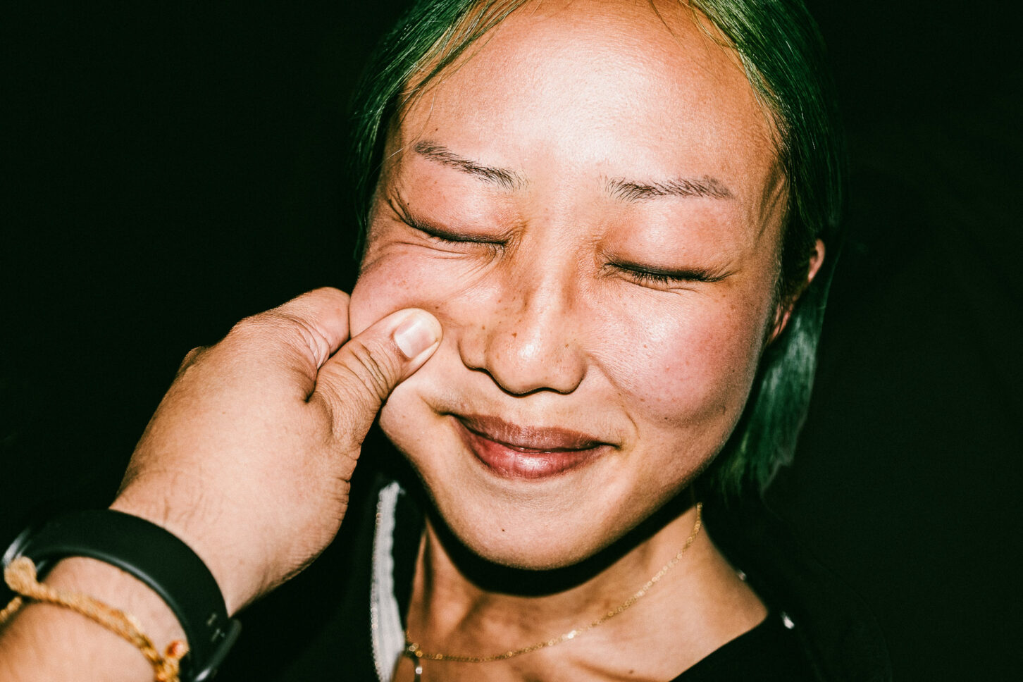 A woman with a playful, annoyed grimace captured with harsh flash lighting during a cheek-pinch for the 'Cheeky Bastards' series.
