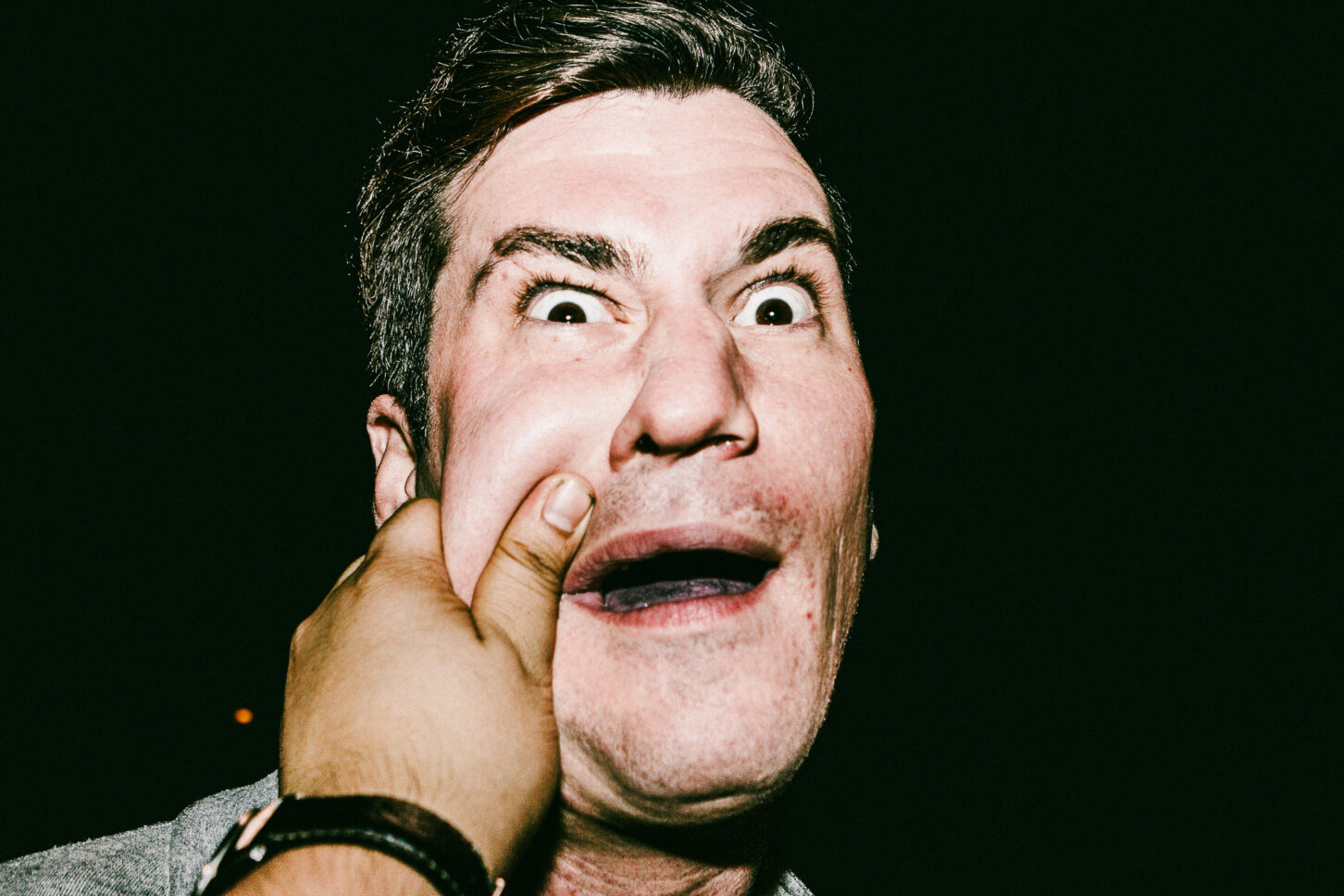 A young man with a furrowed brow and a smirk in a raw flash-lit portrait, showcasing the candid reactions from the 'Cheeky Bastards' project.