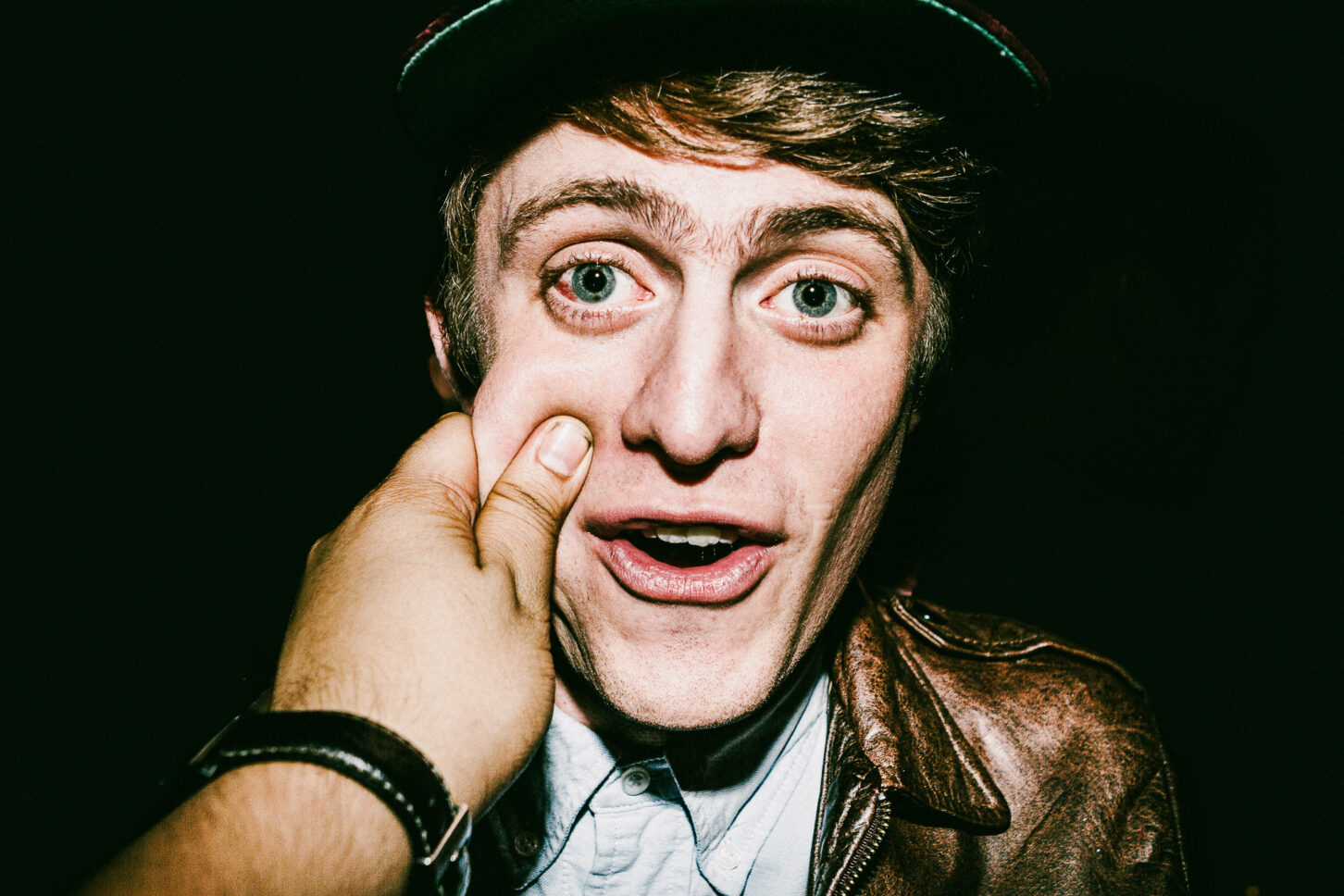 A person with a wide-eyed, shocked expression in a raw, gritty flash portrait from the 'Cheeky Bastards' series, reacting to a pinch on their left cheek.