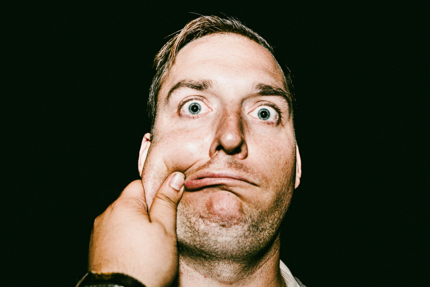 A subject with a confused, furrowed brow in a raw street-style portrait, enduring a left-cheek pinch for the 'Cheeky Bastards' art series.