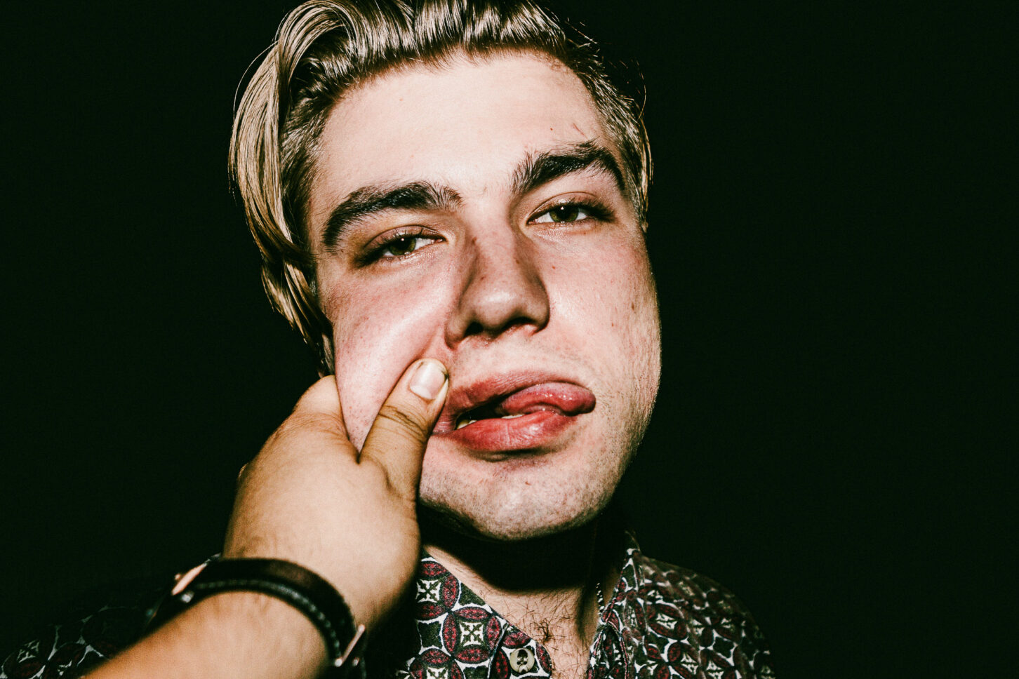 A person looking directly into the lens with an intense, wide-eyed stare while their cheek is pinched, highlighting the raw energy of the 'Cheeky Bastards' series.