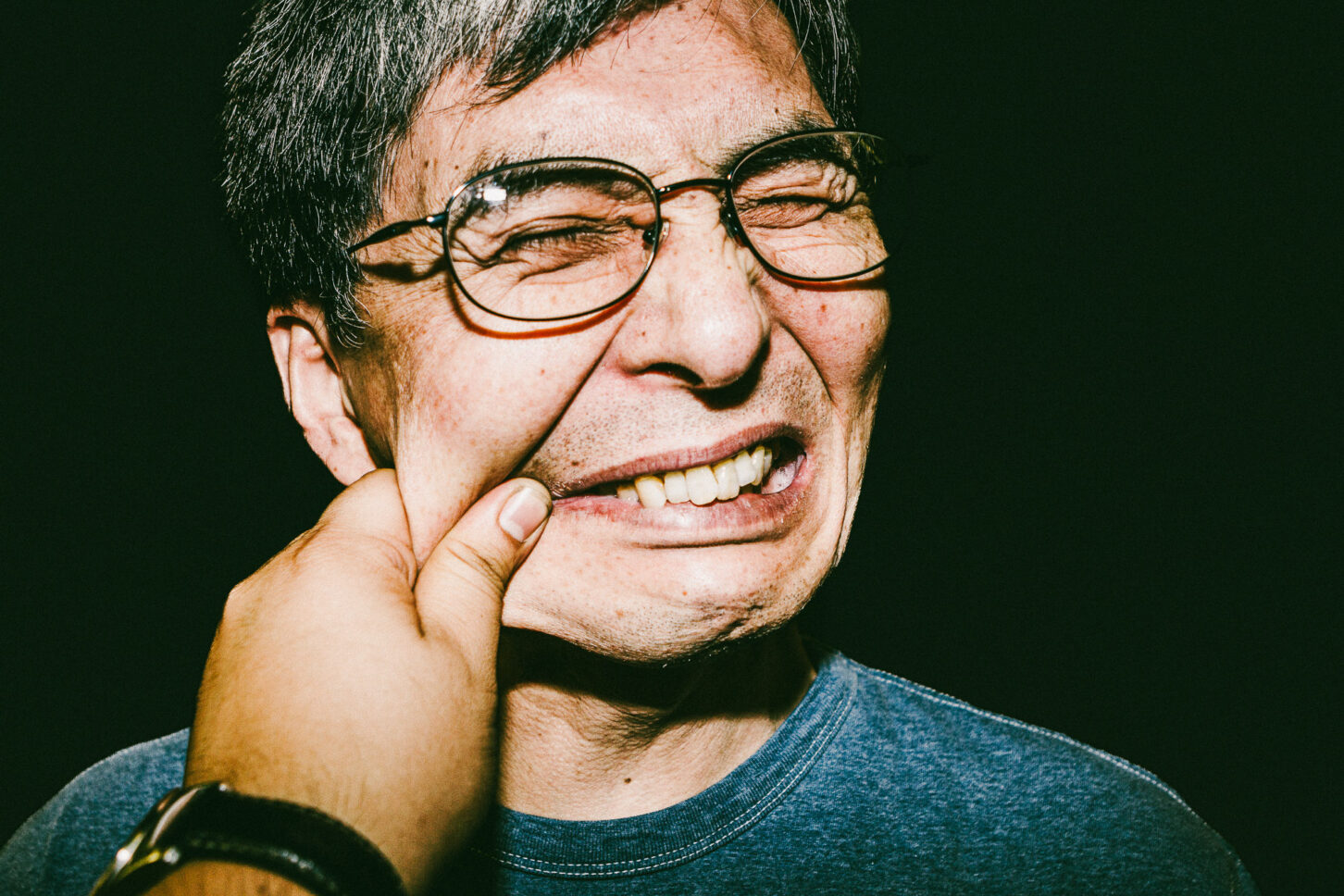 A subject with a wide, toothy grin and squinted eyes, reacting to a cheek pinch in this gritty, low-light flash portrait for 'Cheeky Bastards'.