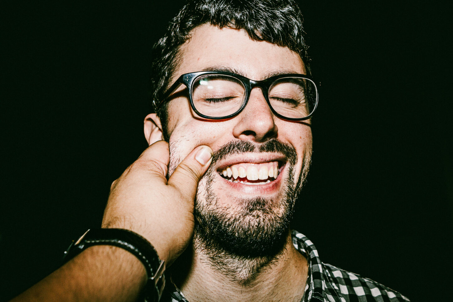 A close-up of a person with glasses adjusting them while reacting to a cheek pinch, captured in the raw style of 'Cheeky Bastards'.