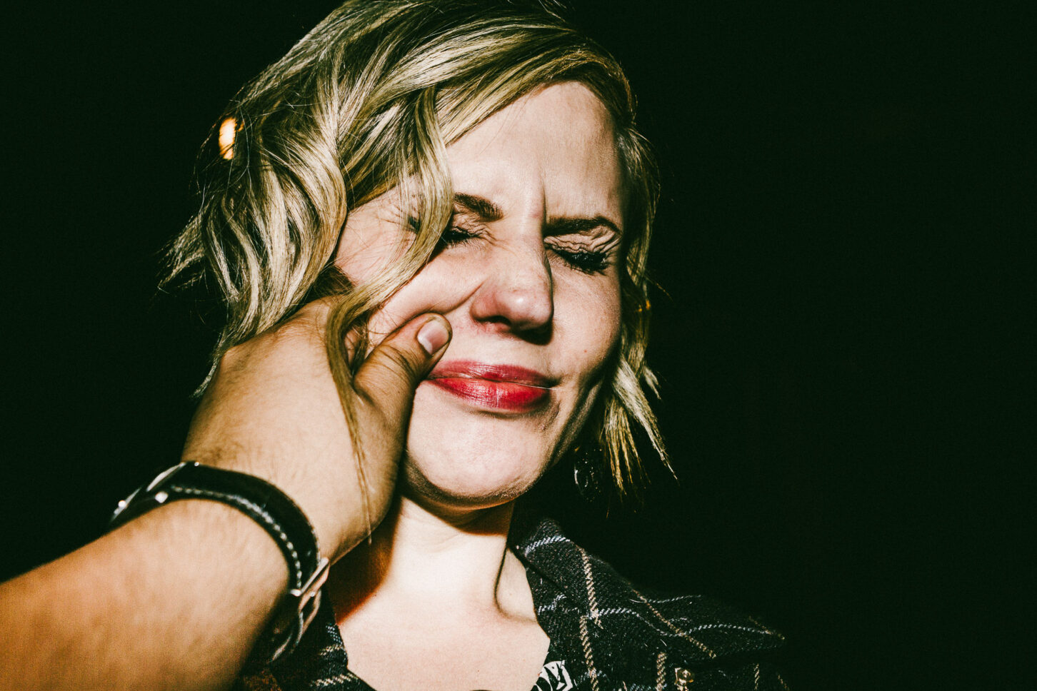 A subject wincing playfully with one eye closed in a raw, high-contrast flash portrait, reacting to a sharp pinch on the left cheek for the 'Cheeky Bastards' series.