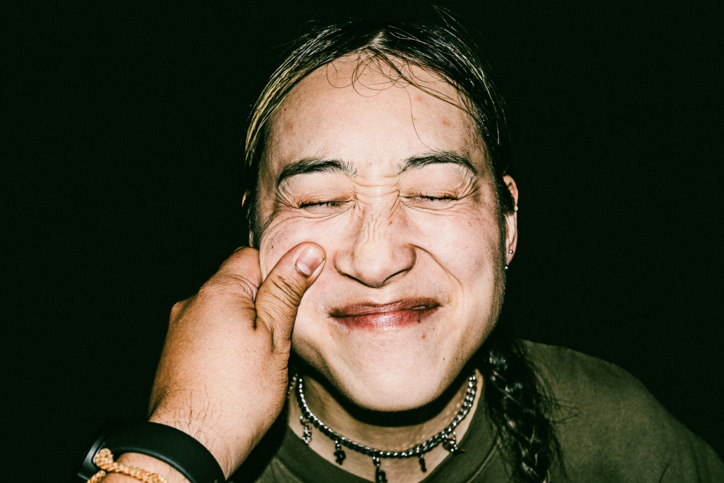 A woman with a playful, annoyed grimace captured with harsh flash lighting during a cheek-pinch for the 'Cheeky Bastards' series.