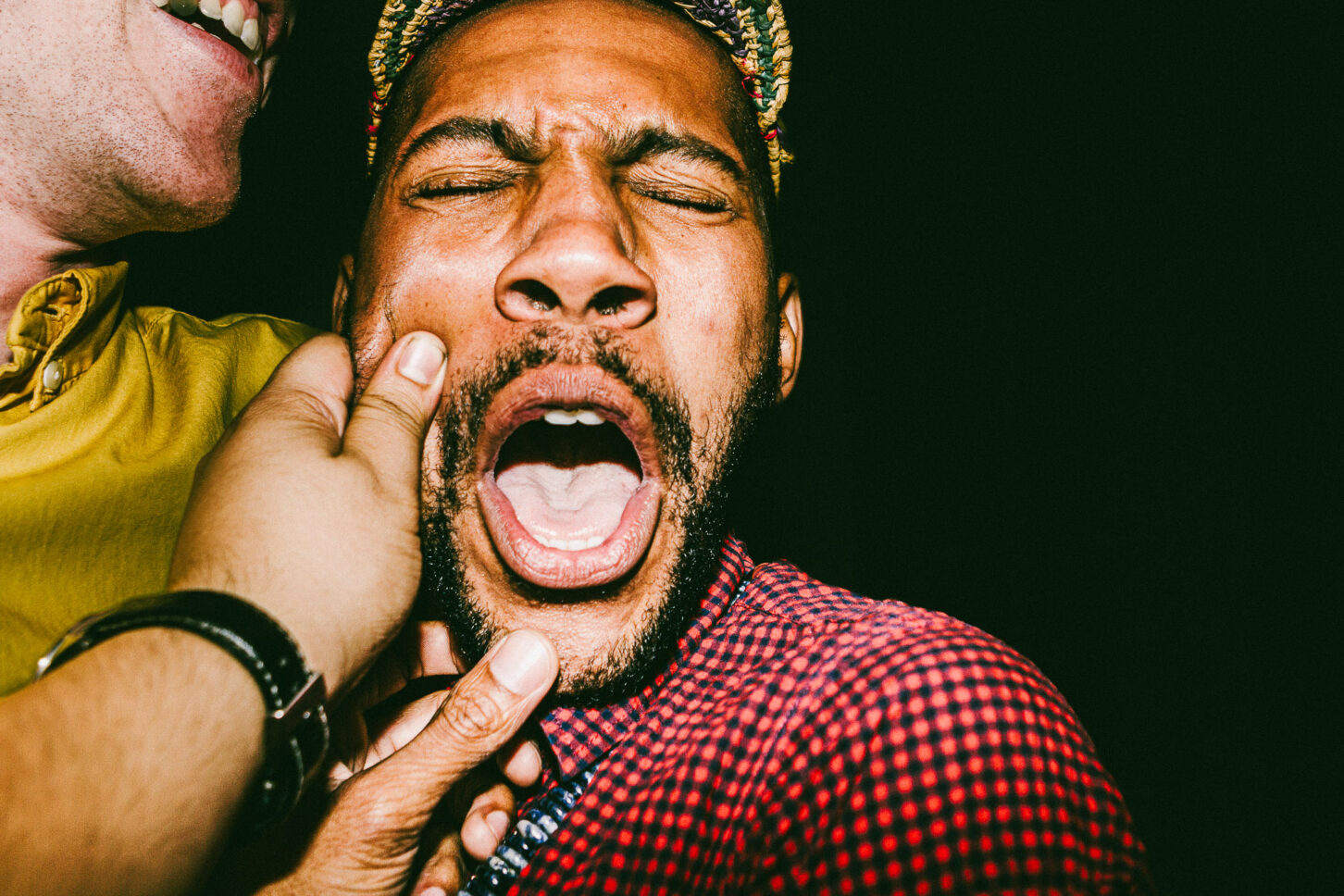 A person mid-laugh with eyes closed in a gritty, high-contrast flash photo, reacting to the signature left-cheek pinch of the 'Cheeky Bastards' series.