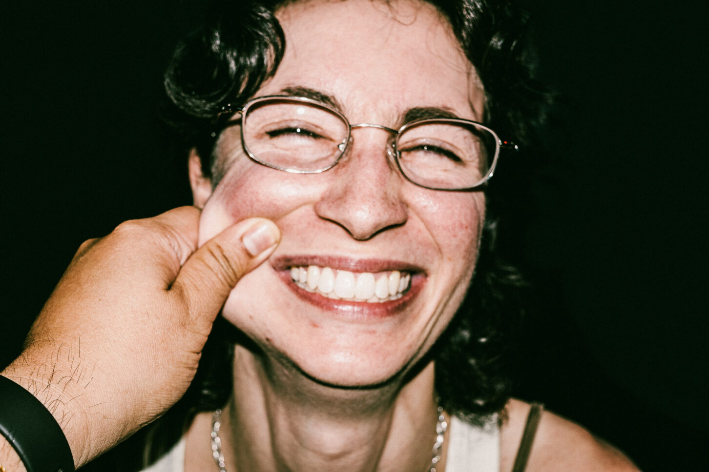 A subject with a wide, toothy grin and squinted eyes, reacting to a cheek pinch in this gritty, low-light flash portrait for 'Cheeky Bastards'.