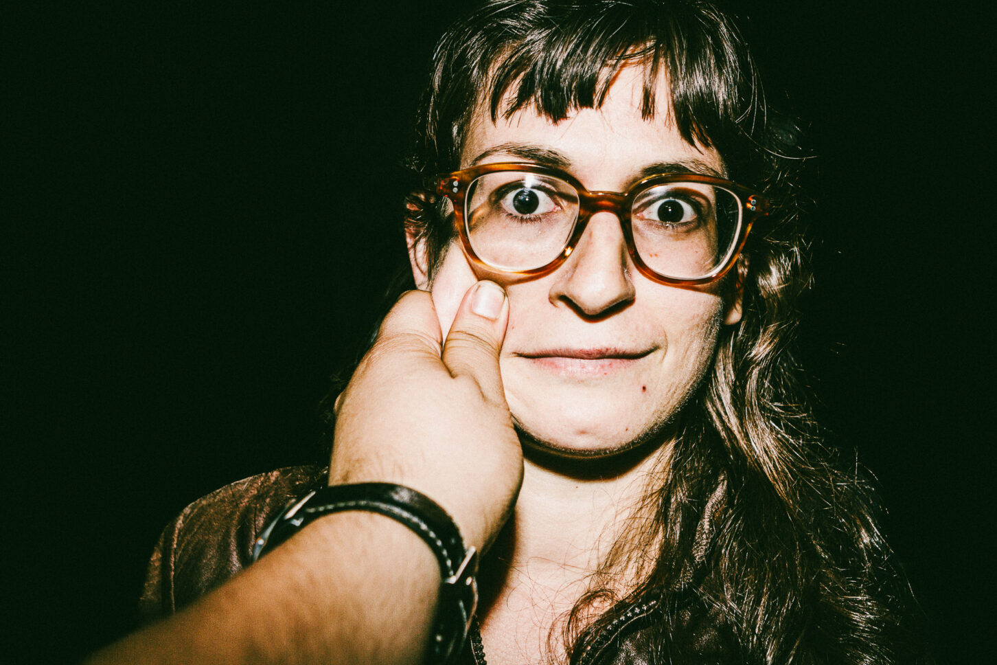 A person looking directly into the lens with an intense, wide-eyed stare while their cheek is pinched, highlighting the raw energy of the 'Cheeky Bastards' series.