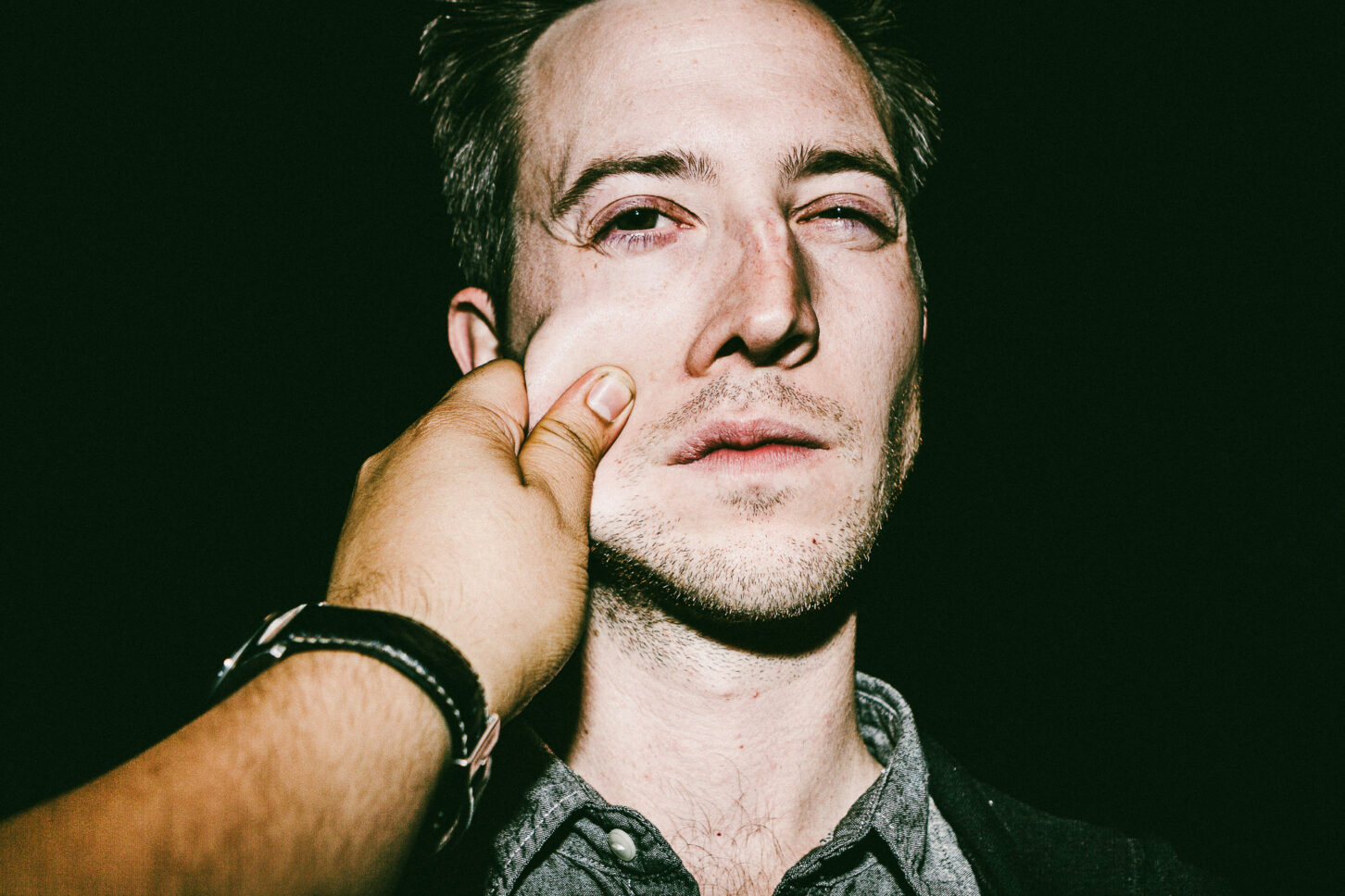 A subject wincing playfully with one eye closed in a raw, high-contrast flash portrait, reacting to a sharp pinch on the left cheek for the 'Cheeky Bastards' series.