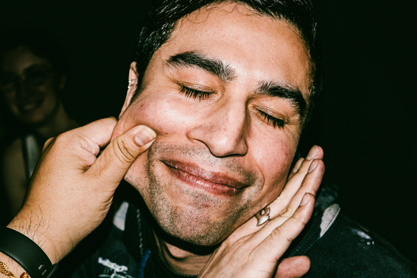 A man with a mischievous, squinted grin in a gritty street-style flash portrait, part of the 'Cheeky Bastards' photography project.