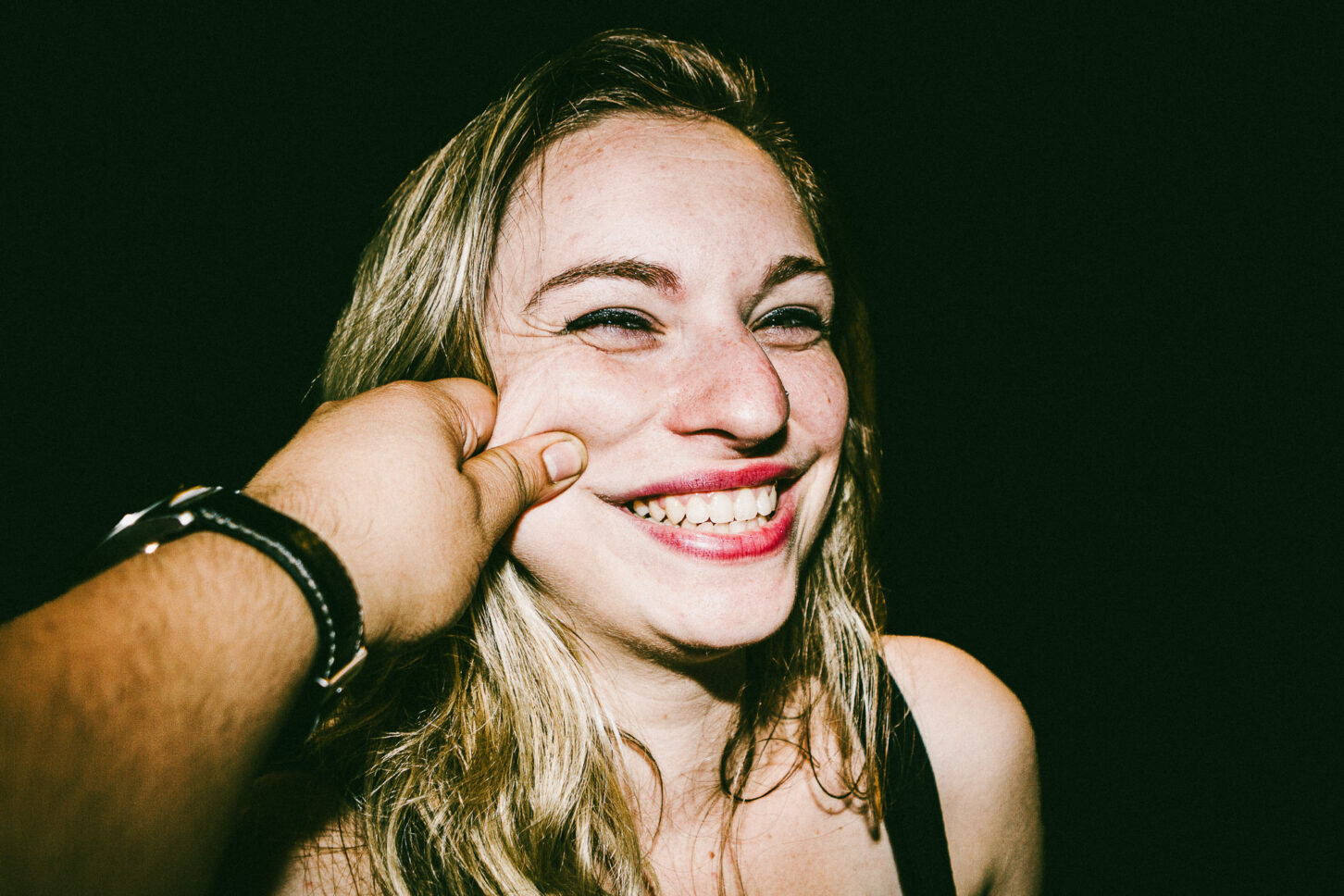 A candid profile shot of a subject turning away while laughing, captured with a harsh flash during a 'Cheeky Bastards' cheek pinch.