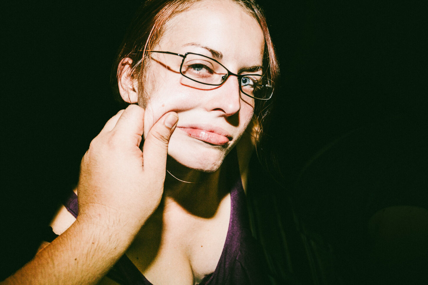 A woman with a playful, annoyed grimace captured with harsh flash lighting during a cheek-pinch for the 'Cheeky Bastards' series.