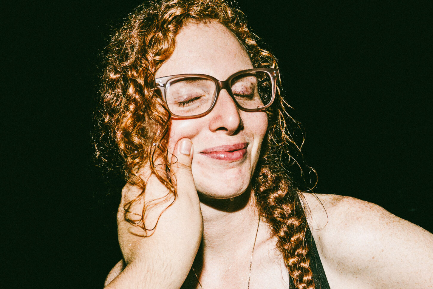 A woman with a playful, annoyed grimace captured with harsh flash lighting during a cheek-pinch for the 'Cheeky Bastards' series.