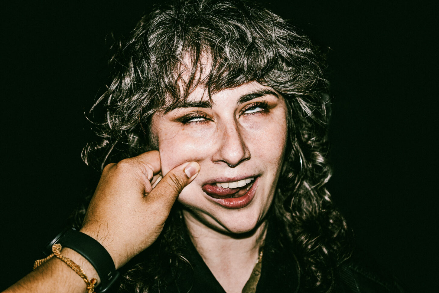 A woman rolling her eyes with a smirk in a candid, harsh-light photo from the 'Cheeky Bastards' series, reacting to the signature cheek pinch.